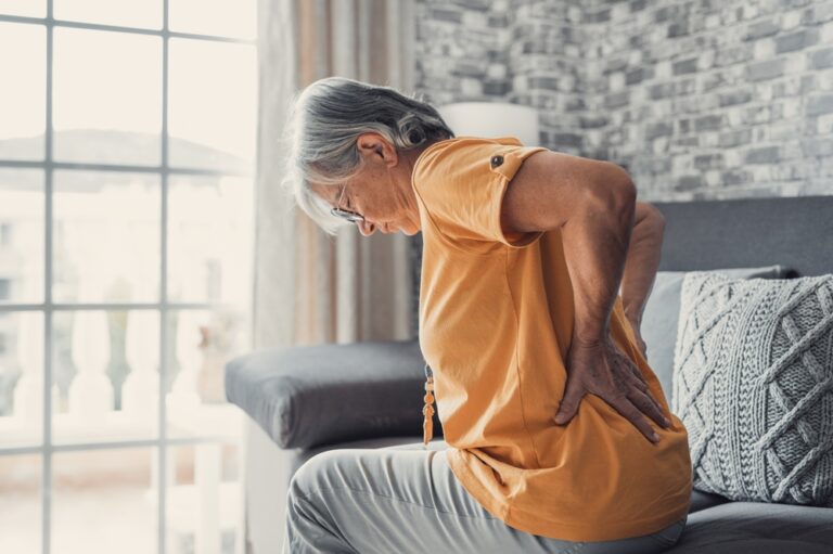 older woman sitting on the edge of her bed, holding her back in pain